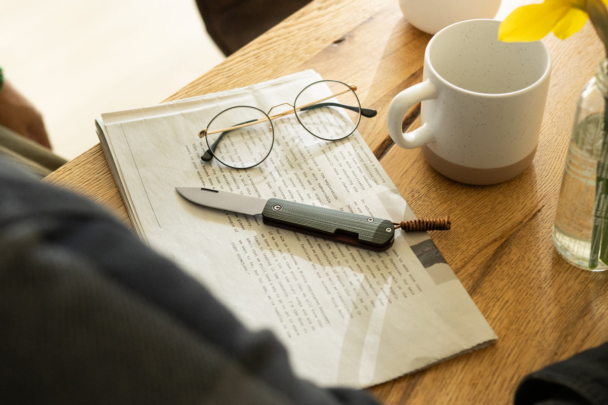 Desk scene with glasses, papers, and a cup beside Slippy Sendy pocket knife