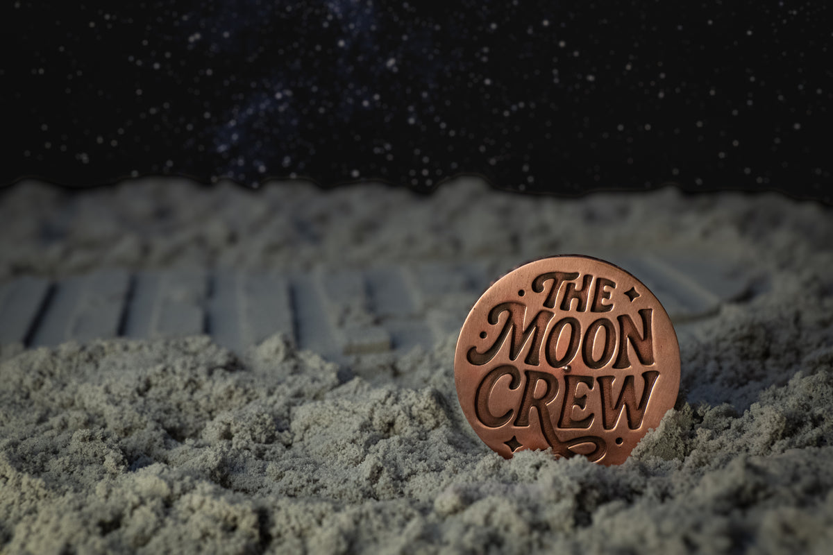 A round copper-colored metal pin or medallion engraved with the words 'THE MOON CREW' stands upright in gray sand under a dark starry sky background.