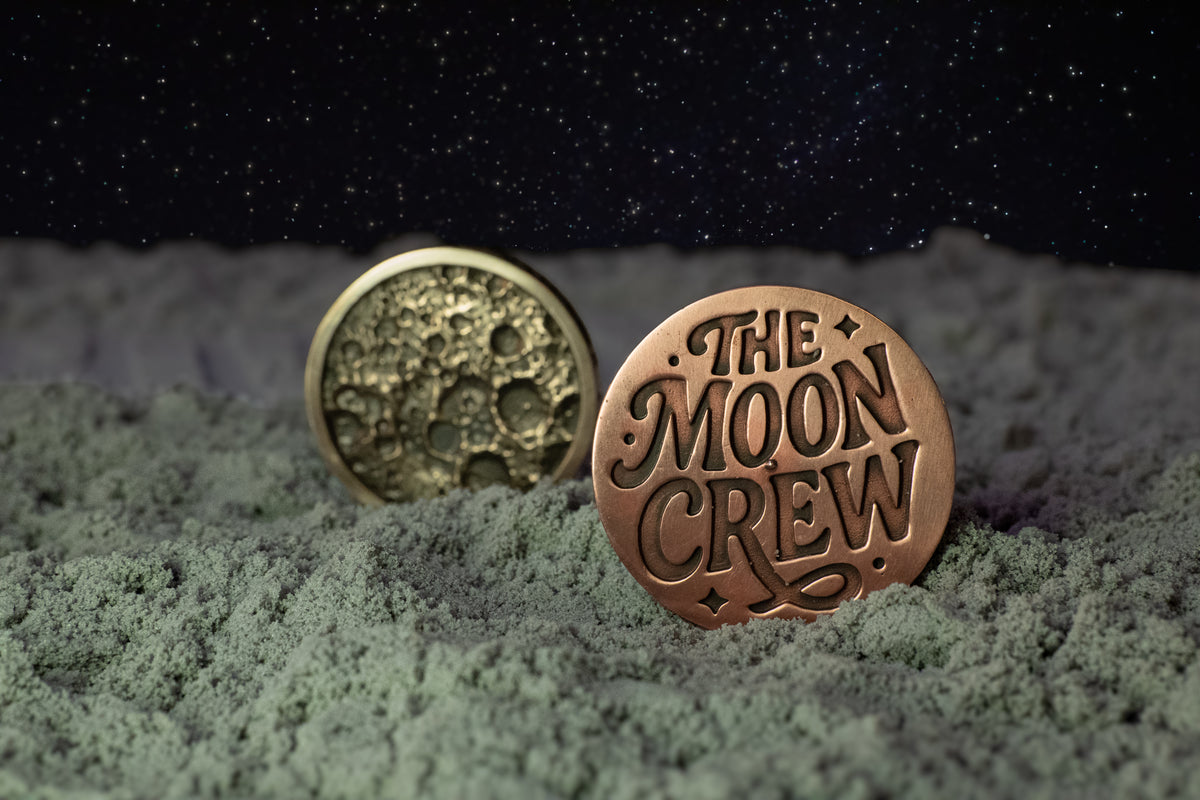 Two round bronze-toned coins or medallions displayed on a textured gray surface under a dark starry sky. One coin is embossed with a crater pattern, and the other reads “THE MOON CREW.