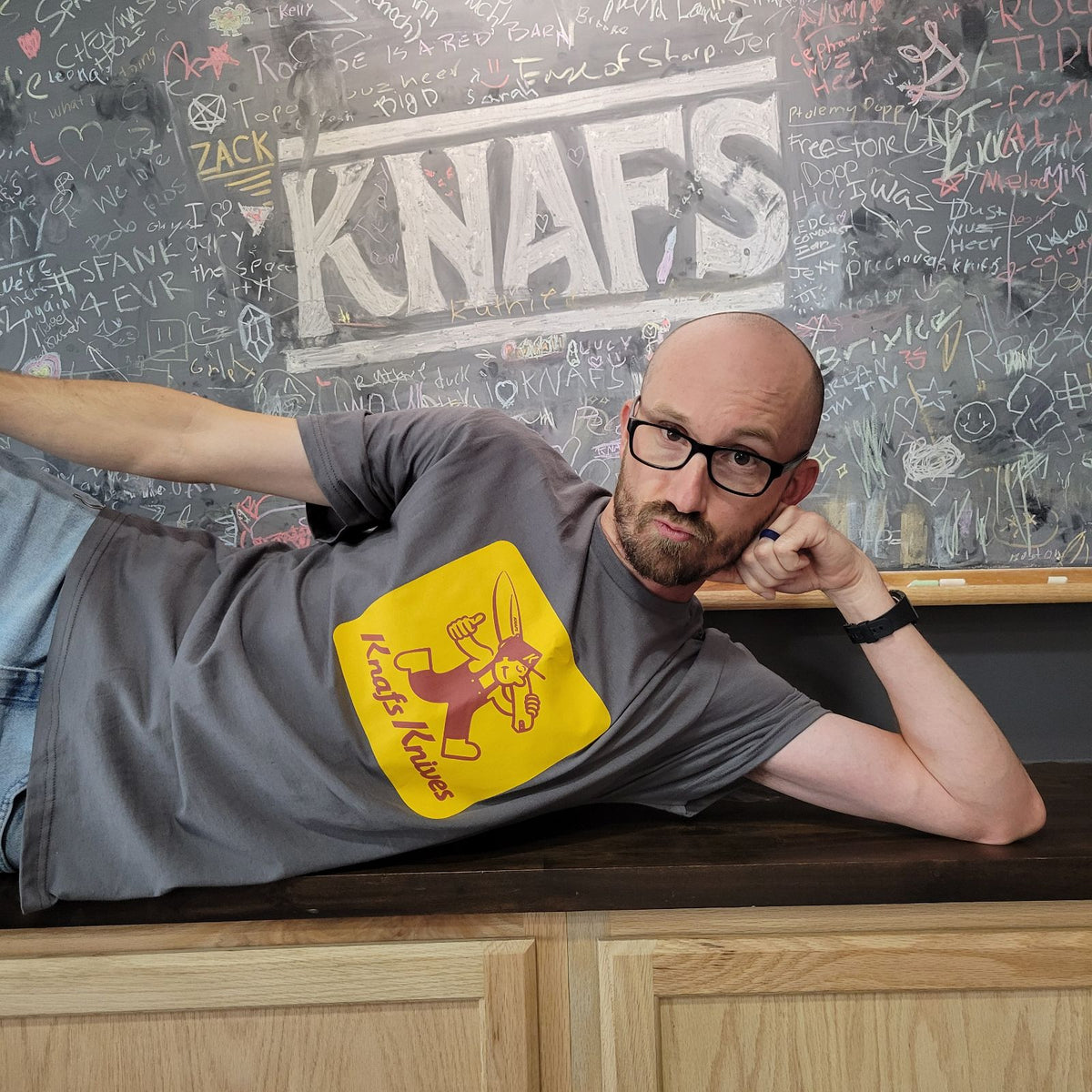 Man leaning on table wearing Knafs Blade Show West 2025 t-shirt with orange graphic