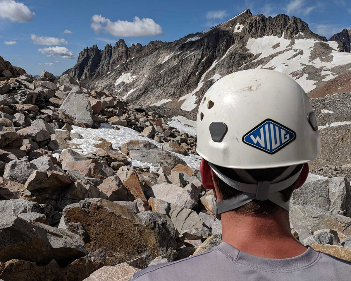 Person wearing a white climbing helmet with a Knafs WIUG sticker, standing on a rocky mountain trail